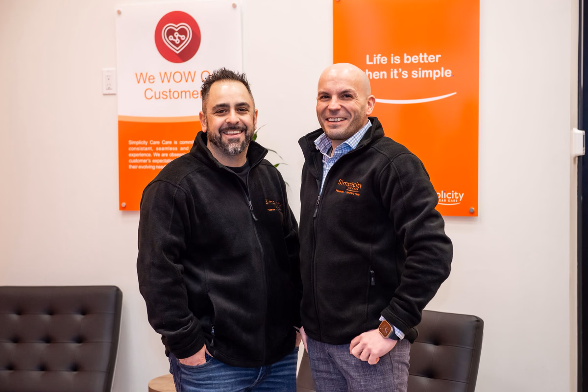 Ultimate Car Care Group owners in front of the Simplicity Car Care values panels: "We WOW Customers" and "Life is better when it's simple."